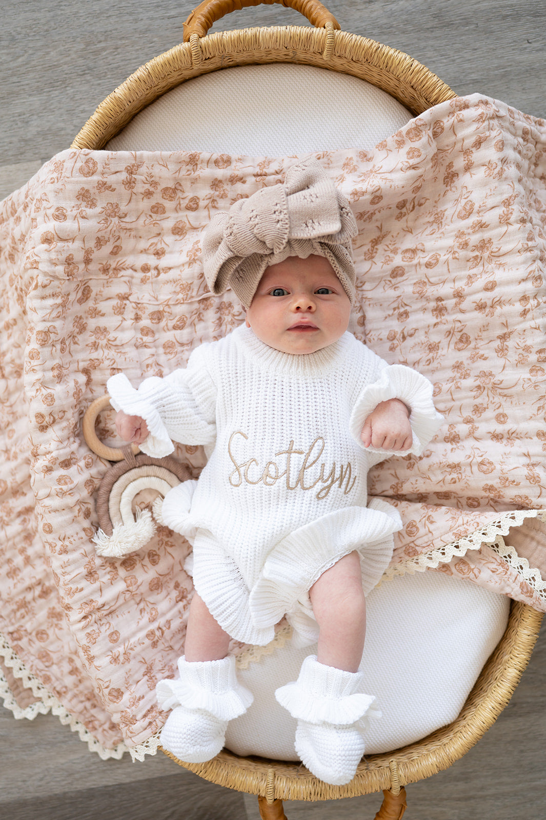 Baby in a white outfit with 'Scotlyn' on it, sitting in a basket with a textured blanket and wooden toys.